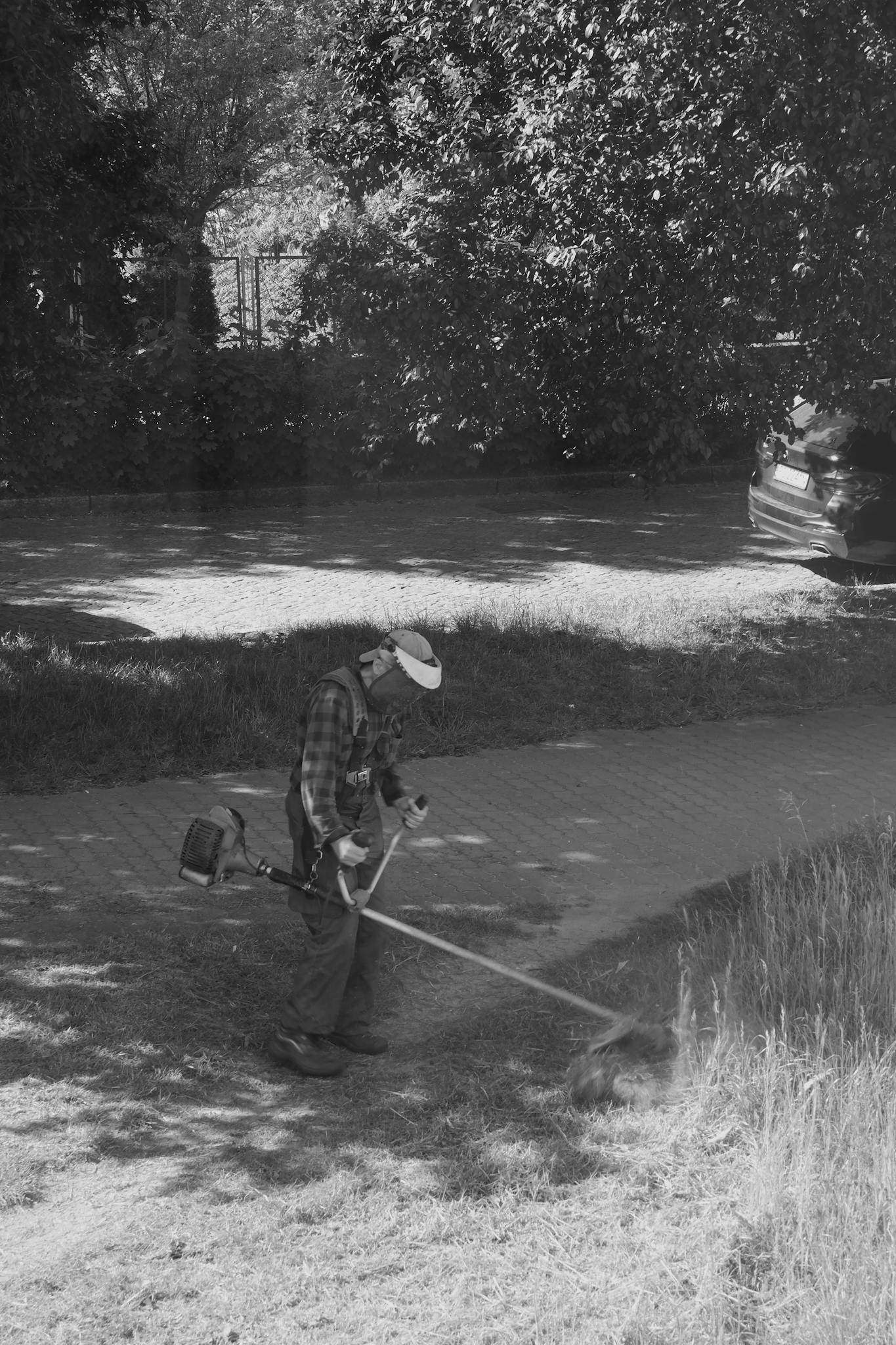 A man wearing a hat is trimming grass with a string trimmer in a shaded outdoor area.